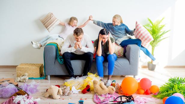 Ein Paar sitzt gestresst auf einem Sofa, während Kinder im Chaos spielen.