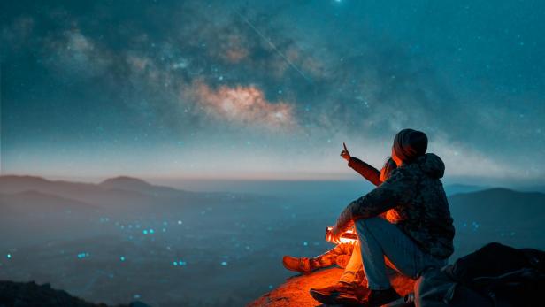Eine Person sitzt auf einem Felsen und blickt in den Sternenhimmel.