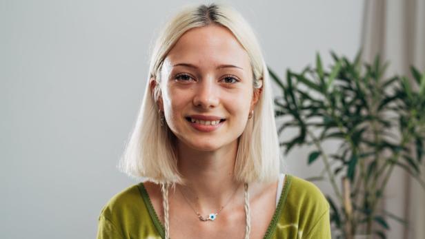 Eine lächelnde junge Frau mit blonden Haaren und einem grünen Oberteil vor einem hellen Hintergrund.