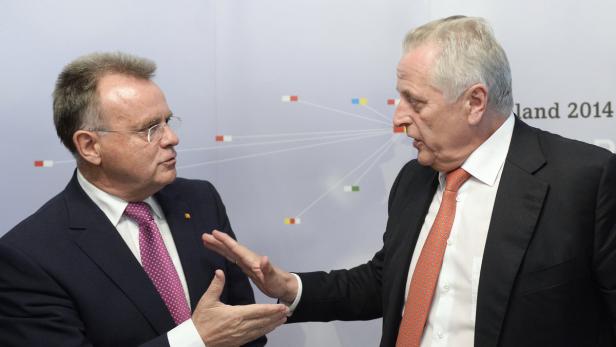 Zwei Männer in Anzügen unterhalten sich angeregt vor einem neutralen Hintergrund.