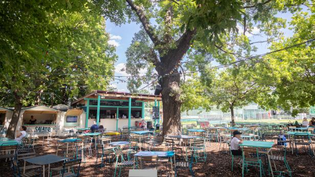 Ein Biergarten mit Tischen und Stühlen unter schattenspendenden Bäumen.