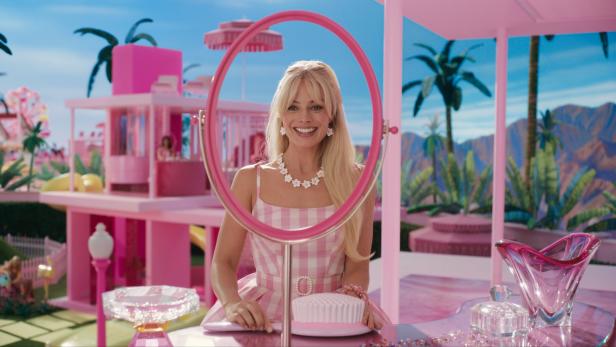 Margot Robbie als Barbie vor ihrem pinkfarbenen Haus mit Pool und Palmen.
