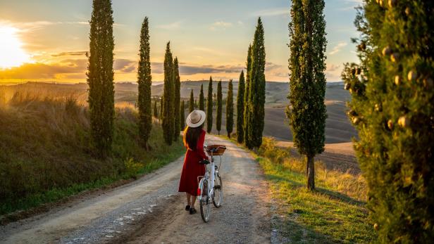 Eine Frau mit Fahrrad geht auf einer Landstraße in der Toskana entlang.