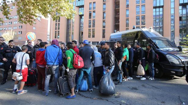 Eine Gruppe von Menschen steht mit Gepäck vor einem Gebäude und einem Polizeiwagen.