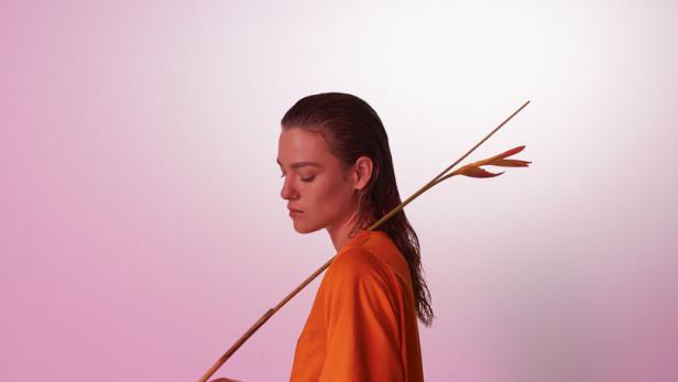 Eine Frau in einem orange-pinkfarbenen Kleid hält eine Helikonienblüte vor einem rosa Hintergrund.