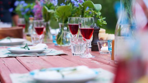 Ein gedeckter Holztisch mit Blumen, Tellern, Gläsern und Getränken für eine Feier im Freien.