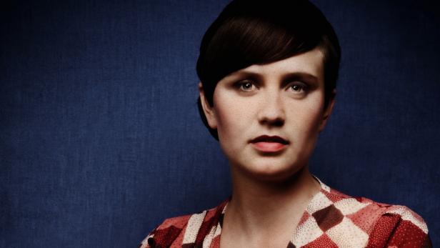 Porträt einer Frau mit braunen Haaren und einem rot-weiß gemusterten Oberteil vor blauem Hintergrund.