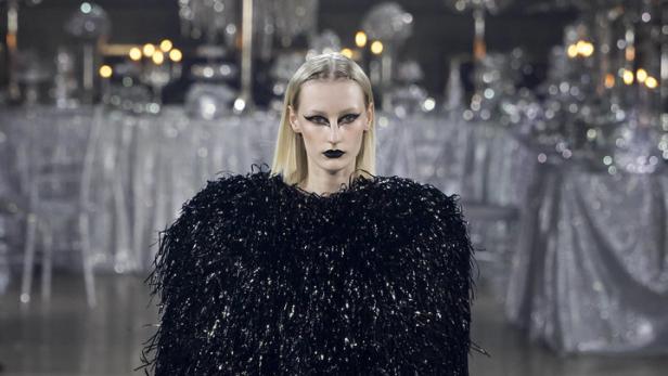 Ein Model mit dunklem Make-up trägt ein schimmerndes, schwarzes Kleid auf dem Laufsteg.