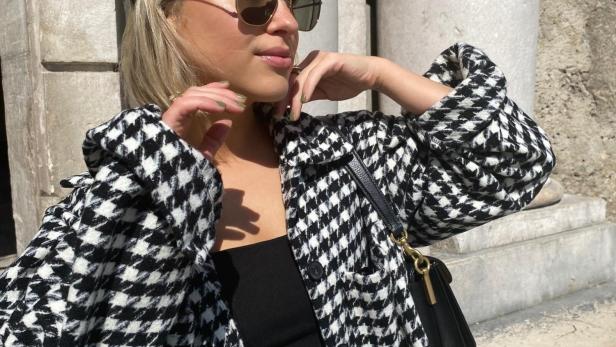 Eine Frau mit Sonnenbrille und Hahnentrittmuster-Jacke posiert vor einem Gebäude.