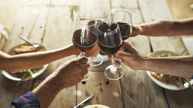 Mehrere Personen stoßen mit Rotwein an einem Holztisch an.