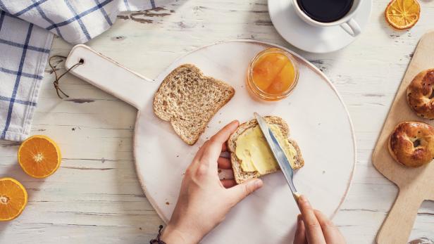 Eine Person bestreicht ein Toastbrot mit Butter, dazu Kaffee und Marmelade.