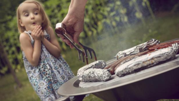 Ein Mann grillt Würstchen und in Folie gewickelte Speisen, während ein kleines Mädchen überrascht zusieht.