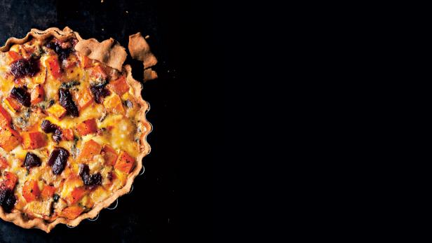 Eine Quiche mit Kürbis, Käse und dunklen Pflaumen auf einem dunklen Untergrund.