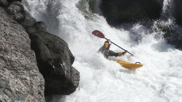 Ein Kajakfahrer kämpft sich durch reißende Stromschnellen neben einem Felsen.