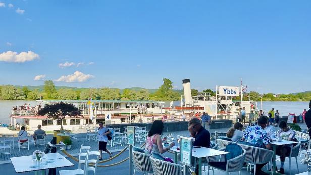 Ein Ausflugsdampfer der Marke Ybbs liegt vor einem Restaurant mit Gästen an Tischen.