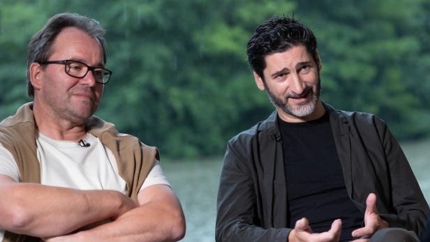 Zwei Männer sitzen vor einem unscharfen Hintergrund und unterhalten sich.