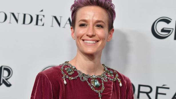 Megan Rapinoe lächelt in einem bordeauxroten Kleid mit Schmuck am Kragen.