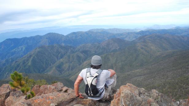 Ein Mann mit Rucksack sitzt auf einem Felsen und blickt auf eine Berglandschaft.