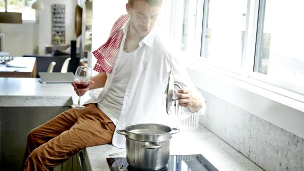 Ein Mann sitzt auf der Küchentheke, hält ein Glas Wein und kocht Pasta.