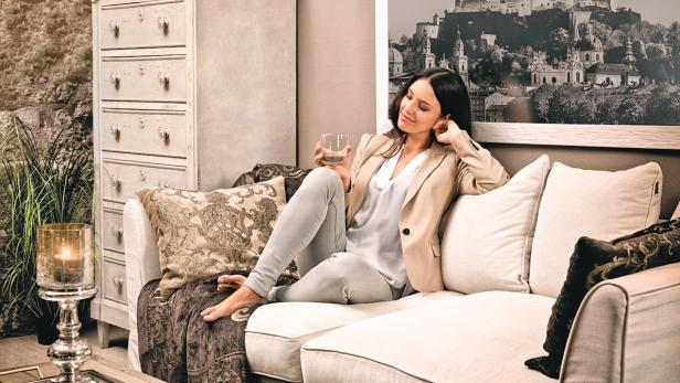 Eine Frau entspannt mit einem Getränk auf einem Sofa; im Hintergrund ein Bild von Salzburg.