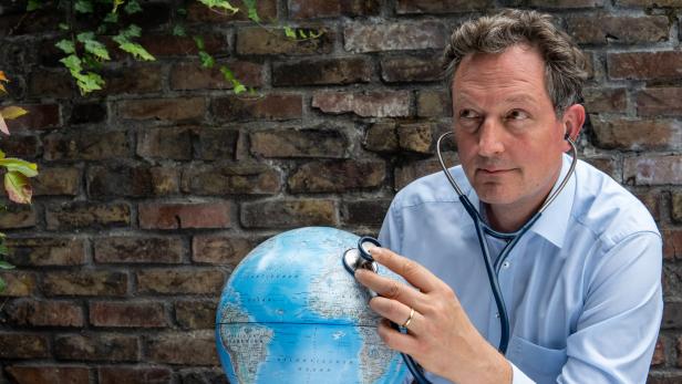 Ein Mann untersucht eine Weltkugel mit einem Stethoskop vor einer Backsteinmauer.