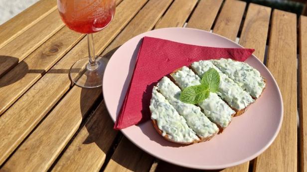 Ein Aperol Spritz und ein Gurkenbrot auf einem Holztisch im Freien.