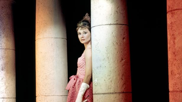 Audrey Hepburn in einem rosa Kleid, posierend zwischen Säulen.