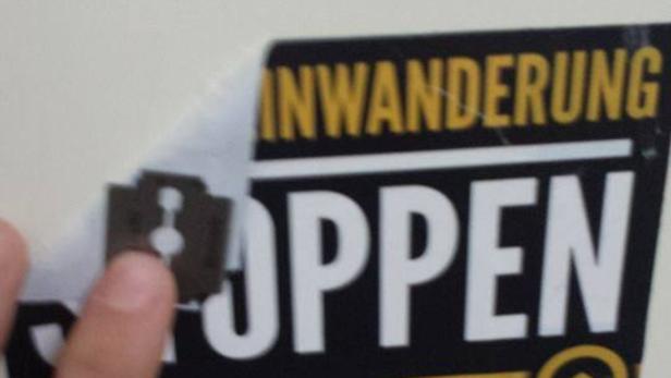 Eine Hand entfernt mit einer Rasierklinge einen Aufkleber mit der Aufschrift „Einwanderung stoppen“.