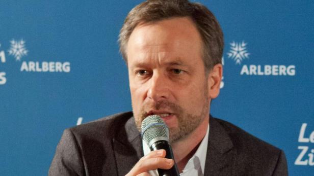 Ein Mann spricht in ein Mikrofon vor einem blauen Hintergrund mit der Aufschrift „Arlberg“.