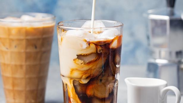 In ein Glas mit Eiswürfeln und Kaffee wird Milch gegossen.