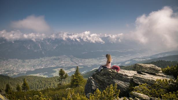 Eine Frau sitzt mit einem Rucksack auf einem Felsen und blickt auf eine Berglandschaft.