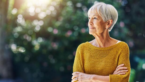 Eine ältere Dame mit verschränkten Armen blickt nachdenklich zur Seite.