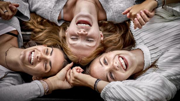 Drei glückliche junge Frauen liegen auf einer Bank und halten Händchen.