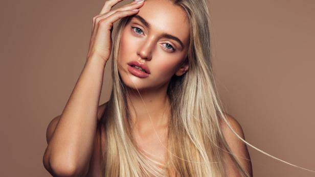 Eine blonde Frau mit blauen Augen posiert vor einem beigen Hintergrund.