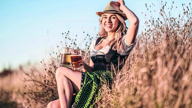 Pretty happy blonde in dirndl, traditional festival dress, sitting with two mugs of beer outdoors in the field