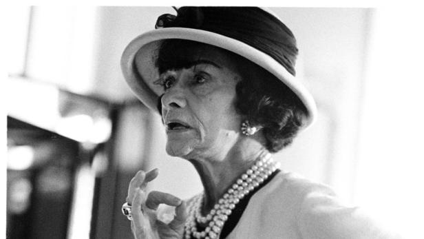 Coco Chanel mit Hut, Perlenkette und Blazer in einer Schwarzweißaufnahme.