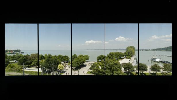 Blick durch ein Fenster auf den Bodensee mit Uferpromenade und Schiffen.
