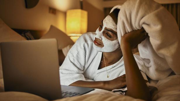 Eine Frau entspannt mit Gesichtsmaske und Laptop im Bett.
