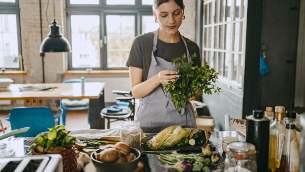 Female chef wearing apron doing quality check of cilantro standing in studio kitchen - Stock-Fotografie