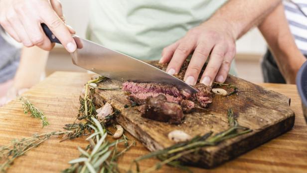 Ein Mann schneidet ein Steak auf einem Holzbrett mit Kräutern und Knoblauch.