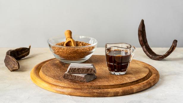 Johannisbrotmelasse, Mehl, Schokolade und Johannisbrotschoten auf Holzschneidebrett mit hellgrauem Hintergrund und Kopierraum - Stock-Fotografie