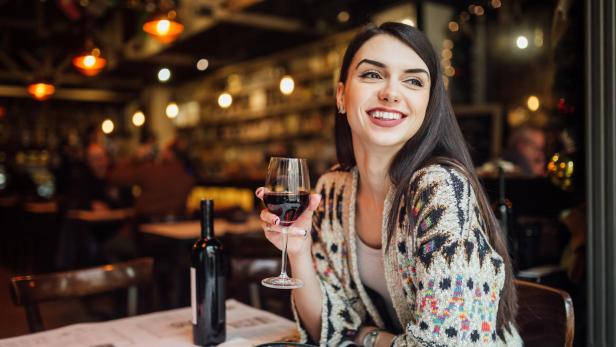 Eine Frau sitzt lächelnd in einem Restaurant mit einem Glas Rotwein.