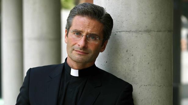 Ein Mann mit Brille und Priesterkragen lehnt an einer Säule.