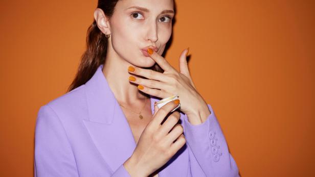 Eine Frau im lila Blazer probiert eine orangefarbene Creme.