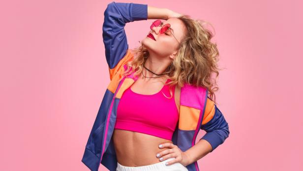 Eine Frau mit Sonnenbrille posiert in Sportkleidung vor einem rosa Hintergrund.