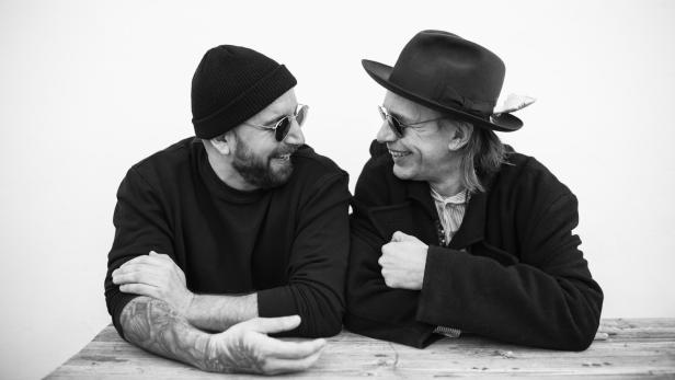 Zwei Männer mit Hüten und Sonnenbrillen sitzen an einem Tisch und schauen sich an.