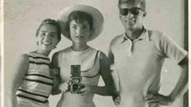 Jackie Kennedy mit einer Kamera, flankiert von einer Frau und einem Mann mit Sonnenbrille.