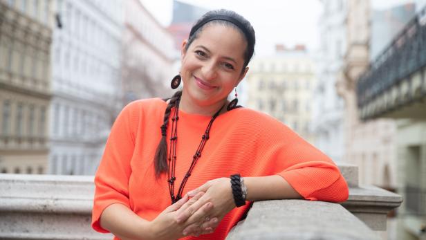 Eine lächelnde Frau in einem orangefarbenen Pullover lehnt an einer Balustrade.