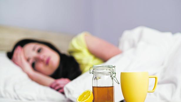 Frau mit Halsschmerzen liegt im Bett und hat die Hausmittel Honig, Zitronen und Tee bereitstehen