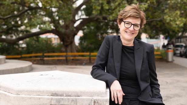 Eine lächelnde Frau mit Brille lehnt an einer Steinmauer in einem Park.